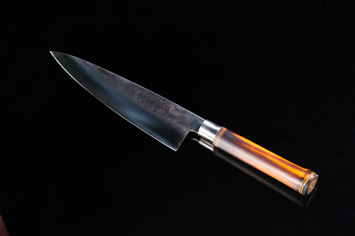 A chef knife with a bamboo handle and a blade that has a dark finish, possibly a high carbon steel or a stainless steel alloy. The handle is partially visible, showing a bamboo texture, and there is a brown wooden sheath present.